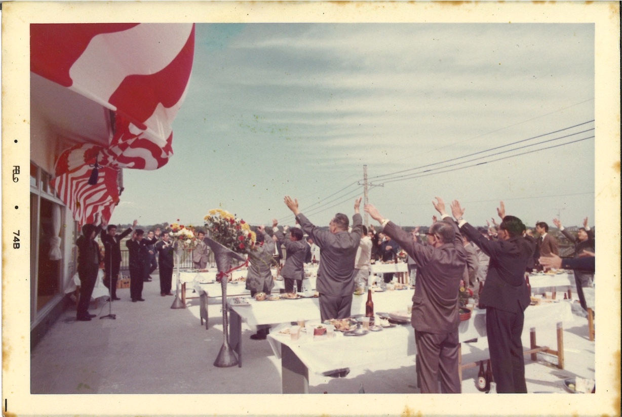 1976年の工場落成式。紅白幕の下で参列者が万歳をしている。